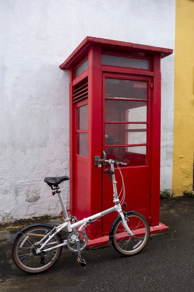 Un vélo pliant blanc garé contre une cabine téléphonique rouge . — Photo