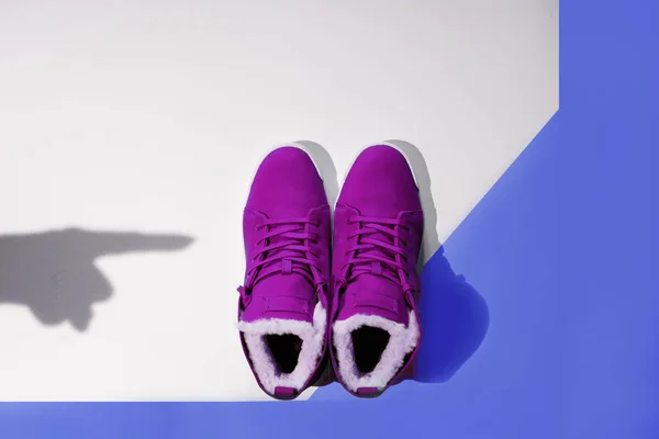 Un par de zapatillas de invierno de color púrpura hembra se encuentra en un fondo azul y blanco con una sombra de una mano femenina que apunta a comprar, flatlay, copyspace, vista superior, mínima, arte —  Fotos de Stock