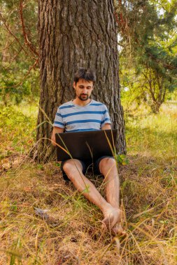 Sakallı genç bir erkek çimenlerde çıplak ayakla oturur ve dizüstü bilgisayara bakar.