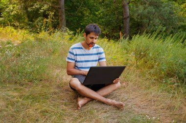 Sakallı genç bir erkek çimenlerde çıplak ayakla oturur ve dizüstü bilgisayara bakar.