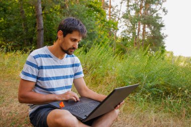 Sakallı genç bir erkek çimenlerde çıplak ayakla oturur ve dizüstü bilgisayara bakar.
