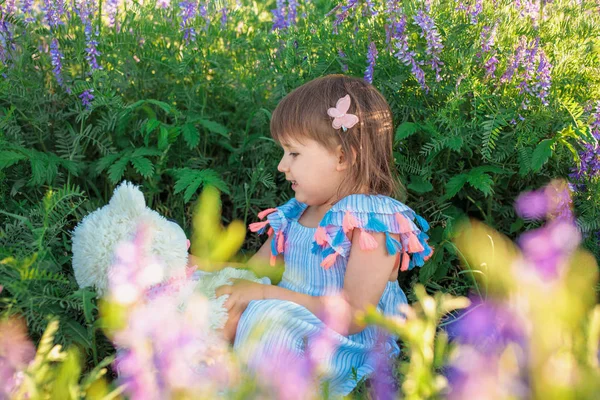 Una bambina di aspetto caucasico siede sull'erba in un abito leggero guarda un orsacchiotto bianco. Senti l'amore e la cura. Concetto di amicizia familiare . — Foto Stock