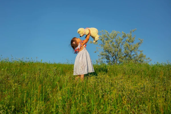 Uma criança de aparência caucasiana fica na grama em um vestido leve, olha para um urso de pelúcia, levantando os braços para o céu. Sinta amor e cuidado. Conceito de Amizade Familiar . — Fotografia de Stock