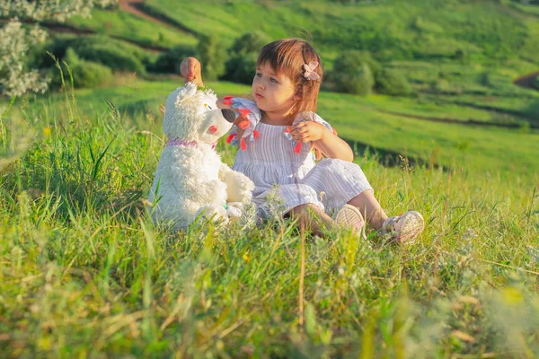 Una bambina di aspetto caucasico siede sull'erba in un abito leggero guarda un orsacchiotto bianco. Senti l'amore e la cura. Concetto di amicizia familiare . — Foto Stock