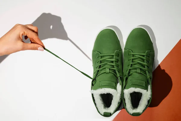 Unisex green winter sneakers on a white and blue background. Female hand pulls shoes by the lace, close-up, flat lay — Stock Photo, Image