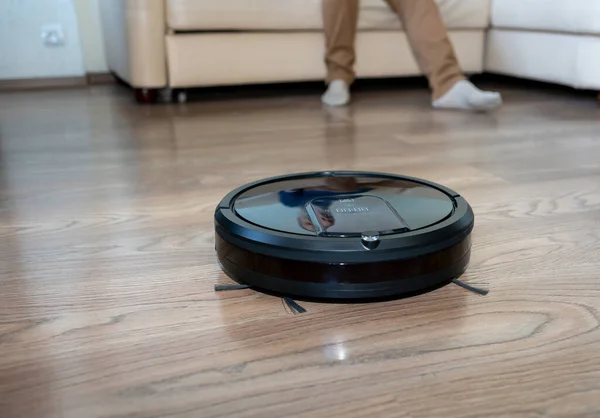Robot vacuum cleaner cleans the floor in the apartment. In the background, you can see the legs of a man sitting on the sofa. Selective focus.