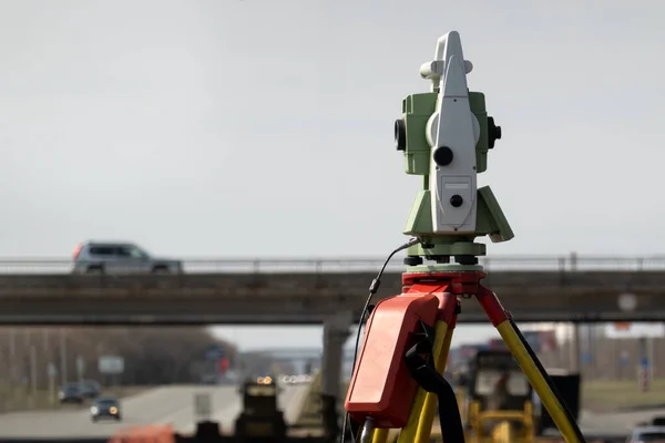 Geodetic support of road construction. In the foreground, a total station on a tripod, in the background, a city landscape and construction equipment. Selective focus.