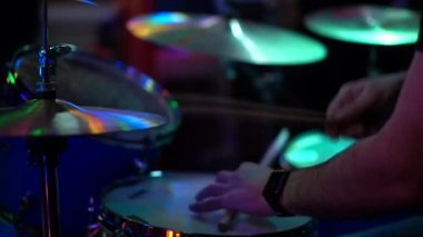 Erkek bir davulcu davul setinin arkasında oturur ve bagetle Hi-hat zilini çalar. Yavaş çekim, yakın çekim, gece kulübünde yapay ışıklandırma.