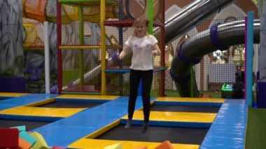 Beyaz tişörtlü ve siyah pantolonlu bir kadın trambolinde zıplıyor, sonra renkli köpük blokları olan bir çukura atlıyor ve ağır çekimde gülüyor.