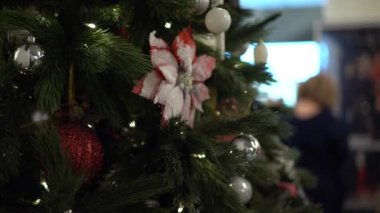 Arka planda gümüş ve kırmızı balonlarla süslenmiş Noel ağacı, kırmızı karla kaplı çiçekler, yakın plan, bulanık insan siluetleri görülüyor