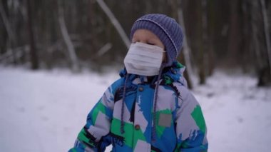 Çocuk (erkek) kışın tulum giyer ve yüzünde tıbbi bir maskeyle kış ormanında şapka standında durur, maske çocuğu engeller ve onu elleriyle sürekli ayarlar.