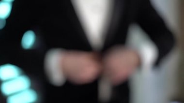 Siyah takım elbiseli bir erkek silueti odak dışı bir şekilde cekete bir düğme takar, kamera yakınlaşır ve odaklanır, bir erkek eli aşağı iner ceketin kenarından tutar.