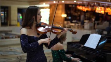 Mavi gece elbisesi giymiş bir kadın keman çalar, ardından yeşil gece elbisesi giymiş bir kadın piyano çalar, klasik konser verir, alışveriş merkezinde 6 ağır çekimde performans sergiler.