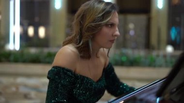 Kahverengi saçlı, Avrupa görünümlü, pullu yeşil bir gece elbisesi giyen bir kadın, yüzün çerçevesinde piyano çalıyor, sonra da tuşlara basan ellerin yakın çekiminde, ağır çekimde.