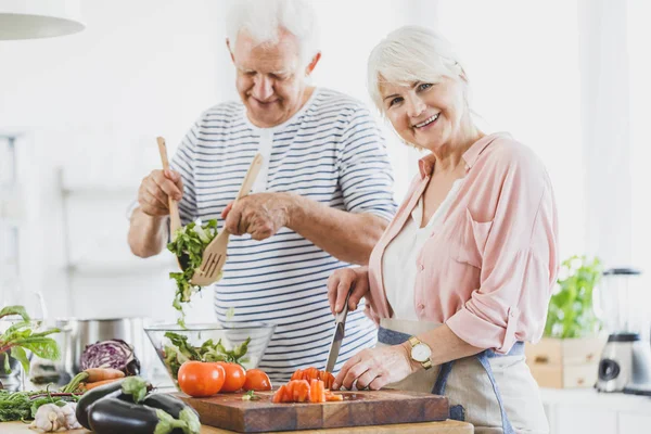 Grandma cuts pepper and grandpa mixes salad — Stock Photo, Image