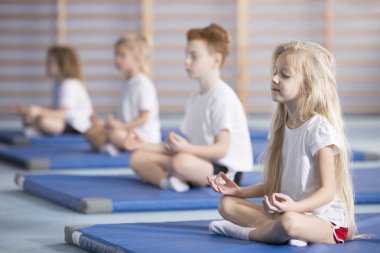 Group of children sitting in lotus pose on blue mats during a yoga class clipart