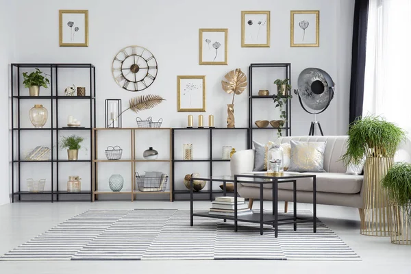 Black table next to couch and plants in living room interior with round gold clock on the wall above shelves