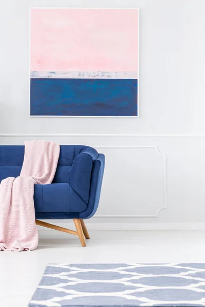 Pink blanket on navy blue sofa against white wall with painting in pastel living room interior