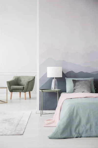 Pink blanket on bed next to a green table with lamp against mountain wall in bedroom interior with armchair