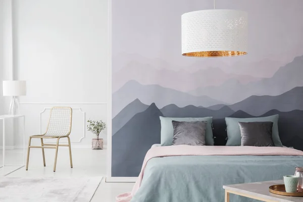 White lamp above pink and green bed in spacious bedroom interior with gold chair and mountain wallpaper
