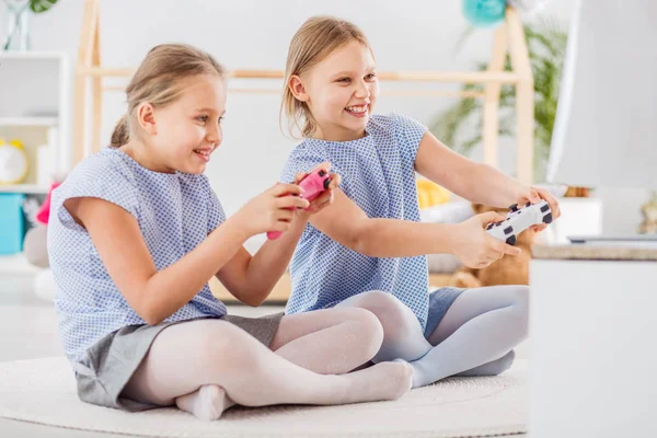 Two girls playing video games sitting on a white rug looking happy and holding pads