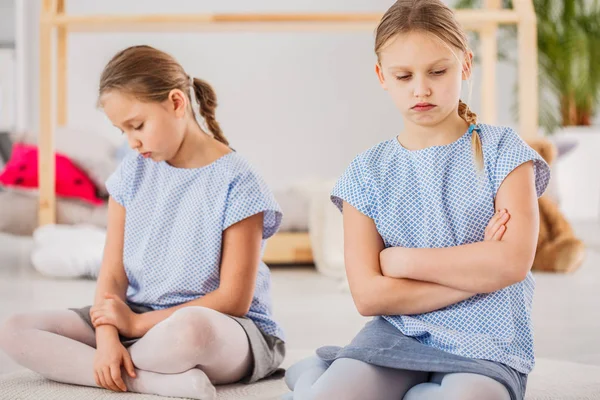 Twin sisters sad after a fight, looking down, sitting with her arms crossed in a bright kid\'s bedroom