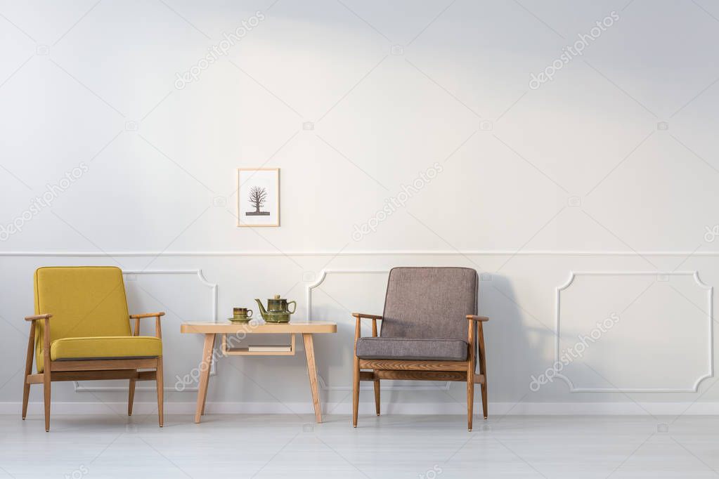 Wooden table between yellow and grey armchair against white wall with poster in living room interior