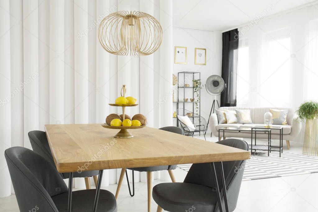 Bright dining room interior with fruit on wooden table, gray chairs and tubes wall. Living room in the background