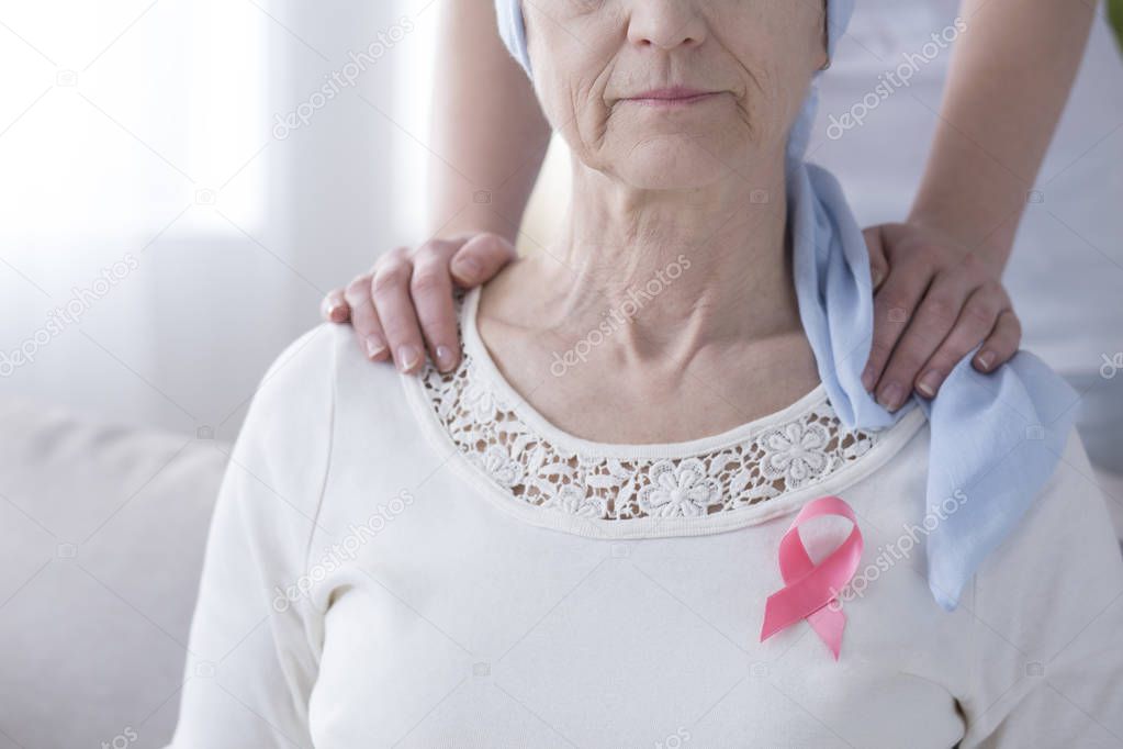 Breast cancer awareness concept - pink ribbon on a sick, older woman and supportive hands on her shoulder