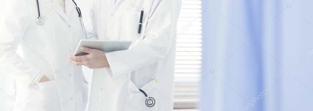 Close-up of a doctor in white coat with stethoscope holding a tablet, consulting on a case with a colleague in bright hospital