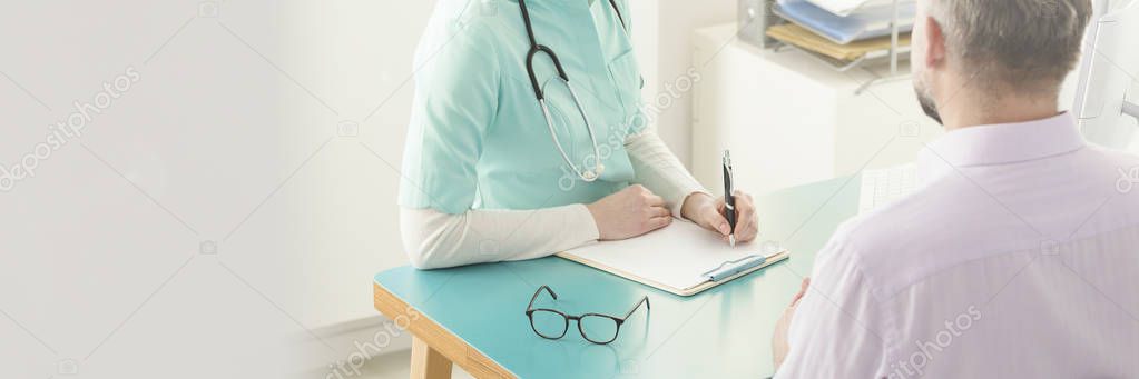 Doctor in green scrubs conducting an interview and writing down patient's medical history information during a consultation