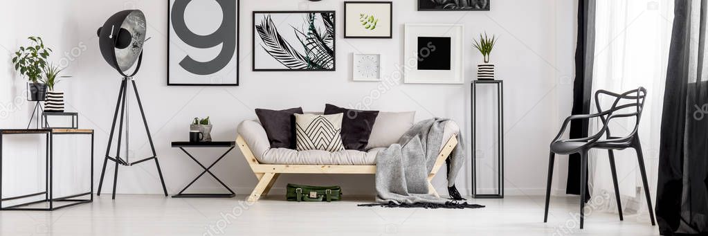 Black big studio lamp placed in the corner of white living room interior with wooden couch, many posters and plastic chair