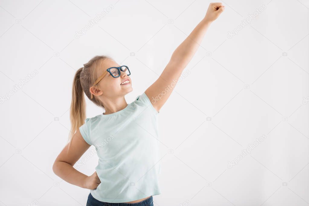 Portrait of a young girl with her arm and fist up and the other arm on a hip in a superhero pose on white wall background