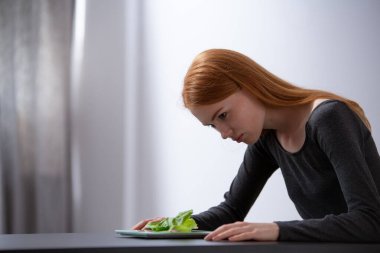 Diyet yapan kızıl saçlı genç kız masada oturuyor ve marullu tabağa bakıyor.