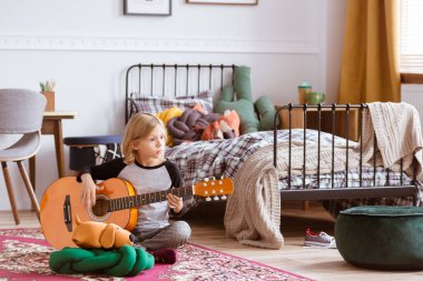 Sevimli sarışın kız son moda odasında yerde oturuyor ve gitar çalmayı öğreniyor.