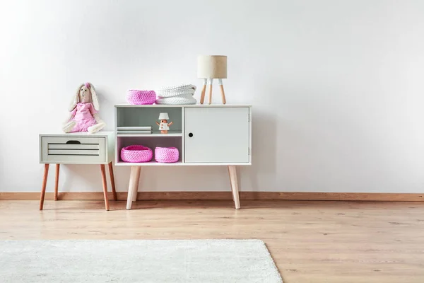 Kleines Spielzeug in pinkfarbener Kleidung auf weißem hölzernen Nachttisch neben weißem Schrank mit elegantem Interieur — Stockfoto