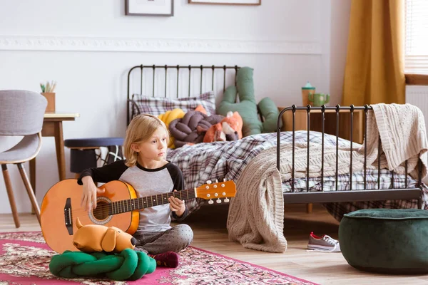 Cute blondynka siedzi na podłodze jej modnej sypialni, uczy się grać na gitarze — Zdjęcie stockowe