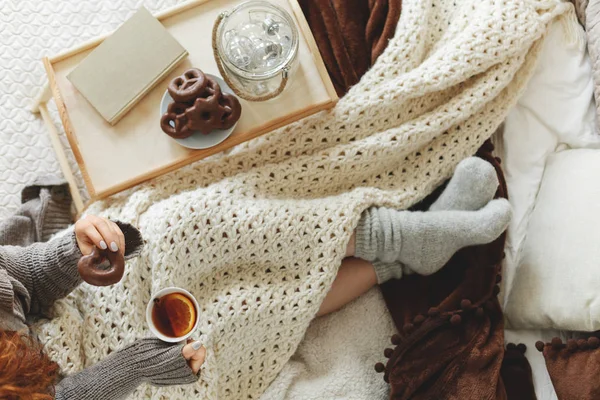 Visão superior da menina coberta com cobertor branco aconchegante sentado na cama beber chá e comer biscoitos — Fotografia de Stock