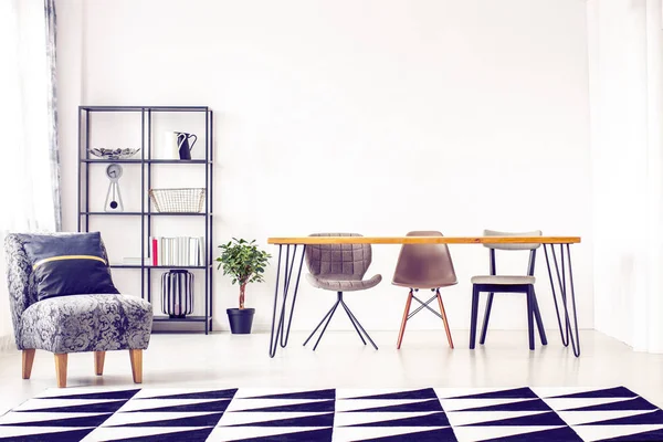 Kussen op grijze fauteuil en geometrisch tapijt in lichte eetkamer met plant op de vloer naast houten tafel met trendy stoelen — Stockfoto