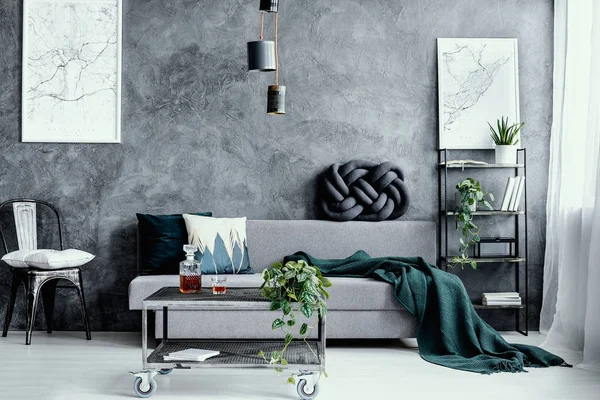 Travesseiro branco na cadeira de metal preto ao lado do sofá cinza com travesseiro no interior da sala de estar escura com mapas na parede de concreto vazio — Fotografia de Stock