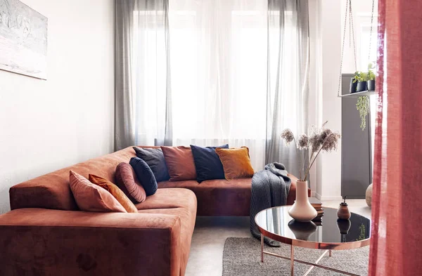 Foto real de travesseiros em um sofá de canto vermelho em pé perto da janela com cortinas cinza no interior da sala de estar — Fotografia de Stock