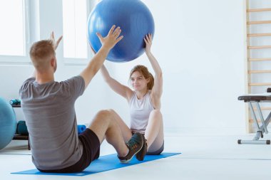 Spor salonunda mavi paspasın üzerinde oturan ve topla antrenman yapan hoş bir hasta.