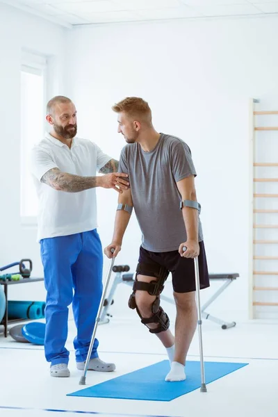 Koltuk değnekleriyle ve ortopedi ameliyatıyla bir adam üstüne bir adam klinikte yürümeyi öğreniyor. Yanında terapist var. — Stok fotoğraf