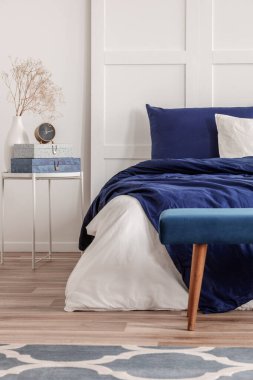 Süslü komodinin üzerinde beyaz vazodaki saat ve kuru çiçekler. Büyük boy yatağın yanında, rahat bir yatak.