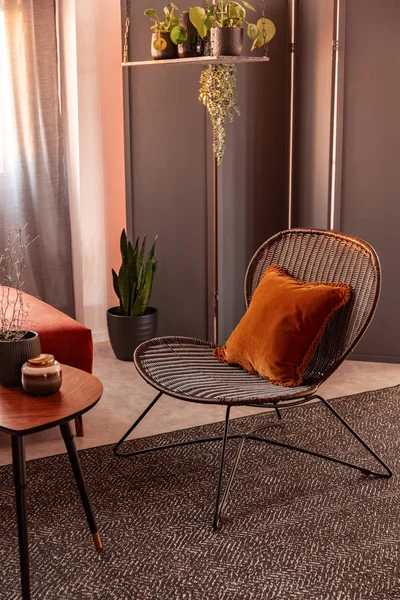 Foto real de um travesseiro laranja em uma cadeira de vime no interior acolhedor da sala de estar com um tapete cinza e mesa de café de madeira — Fotografia de Stock