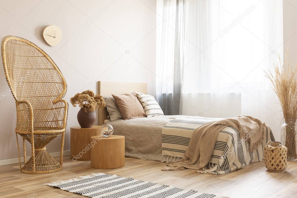 Flowers in brown vase on wooden nightstand in bright bedroom interior with peacock chair