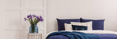 Mor çiçekler mavi cam vazoda, şık komodinin yanında, büyük boy yatağın yanında.
