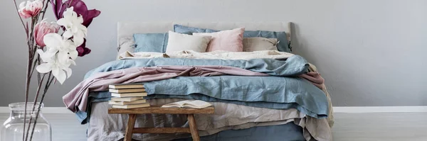 Panoramic view of king size bed with pastel pink, blue and beige bedding next to wooden bench with books, copy space on empty wall — ストック写真
