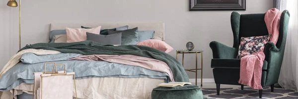 Panoramablick auf King Size-Bett mit pastellrosa, blauen und beigen Betten neben Nachttisch mit Uhr und smaragdgrünem Sessel mit Blumenkissen im stilvollen Schlafzimmerinterieur — Stockfoto