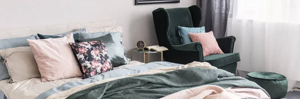 Пастельные розовые и синие подушки на изумрудно-зеленом кресле рядом с тумбочкой с часами и книгой в ярком интерьере спальни с двуспальной кроватью — стоковое фото